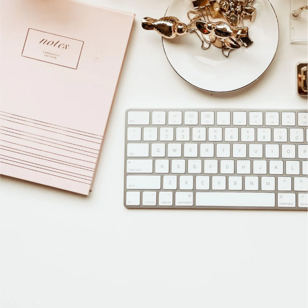 A pink Notes manuscript book, a keyboard and gold stationery on a white desk